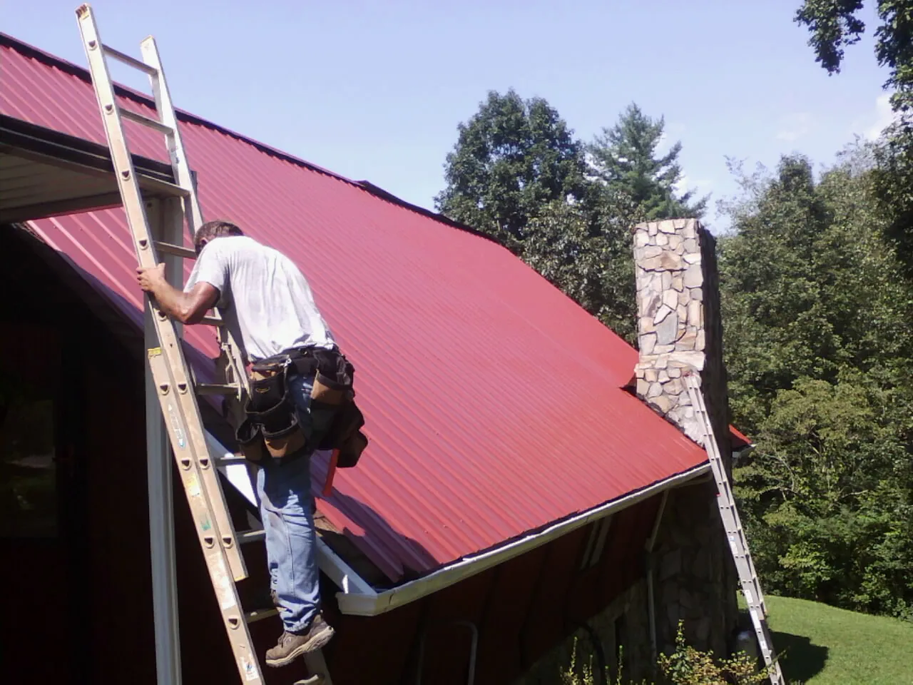 Roofer on a ladder beside a red metal roof and stone chimney — Soffit & Fascia Repair by Roofing Brothers in Lavallette